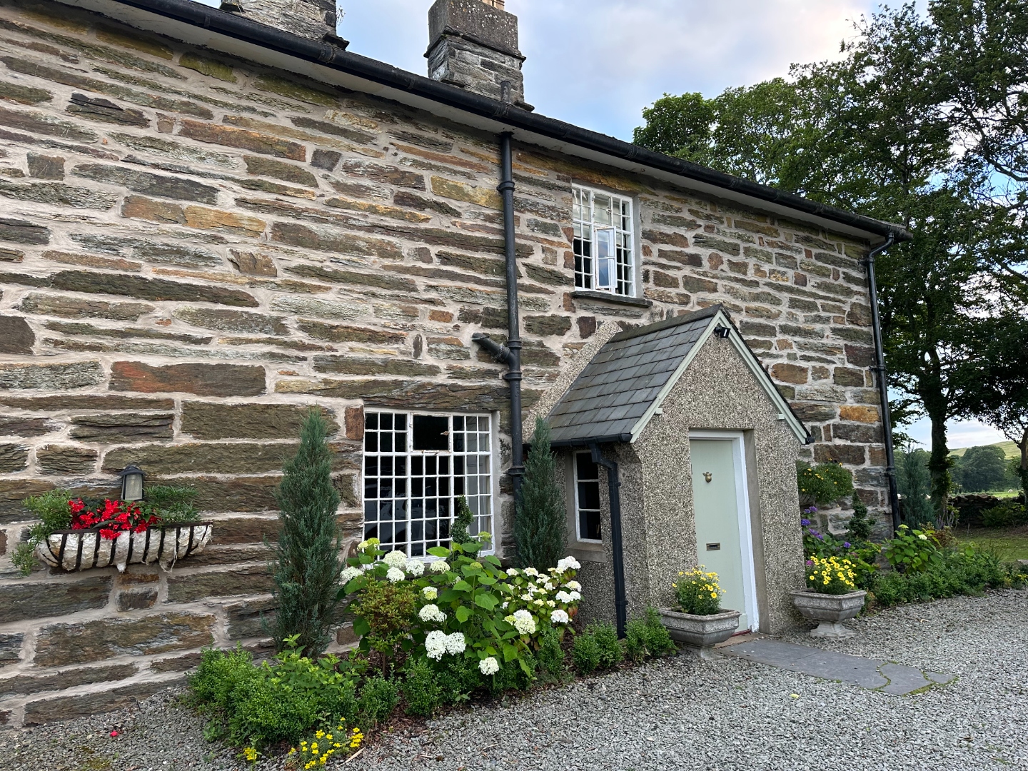 The stone rectory front of Tyddyn Eglwys in summer — hydrangeas in bloom, sage green door, and lush gardens surrounding the Grade II listed building